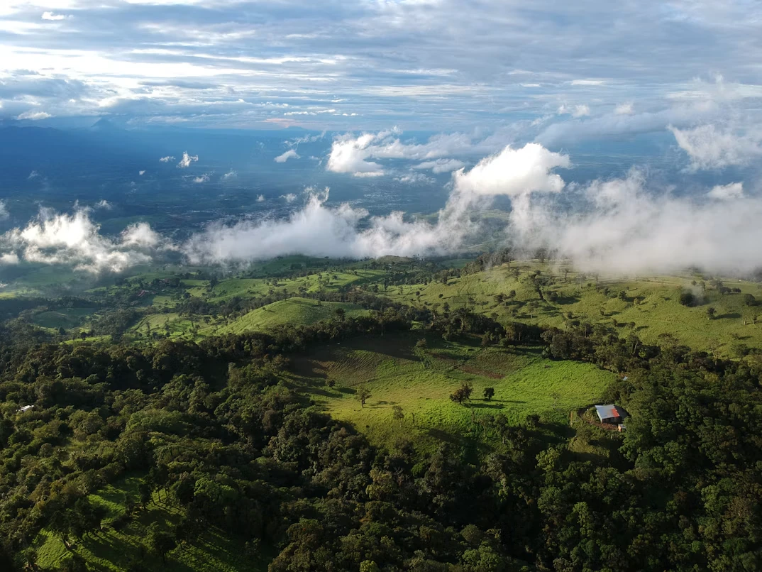 Aerial view over the Costa Rican Central Valley: rolling green pasture and forest with low clouds drifting across the hillsides and a single red-roofed farmhouse.
