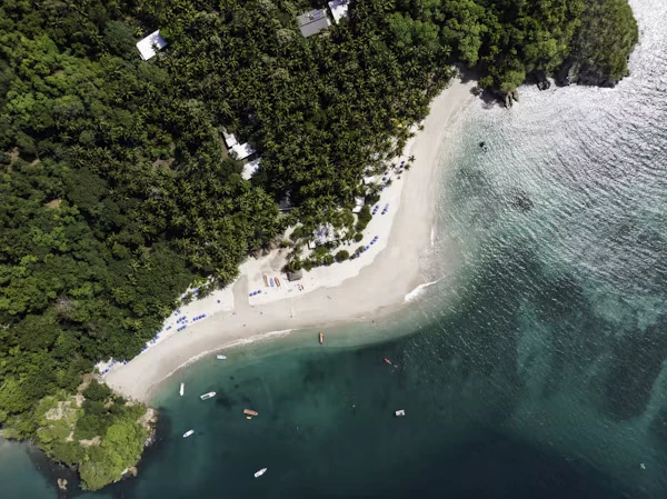 Aerial view of a curved Costa Rican Pacific beach, pale sand edged by dense green jungle, with small boats anchored in clear turquoise water.