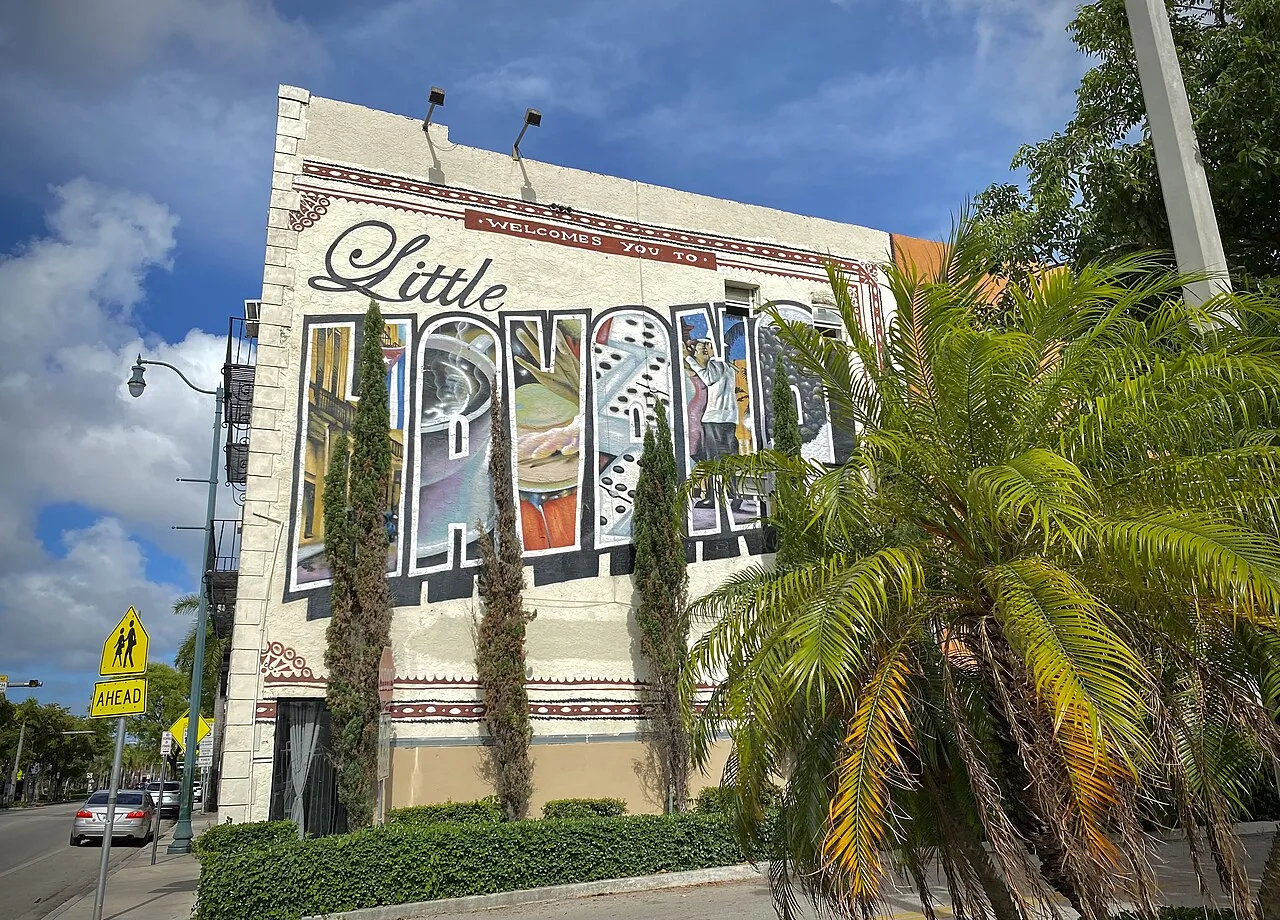 A painted mural on a building reading 'Welcome to Little Havana' in large decorative letters filled with scenes of dominoes, coffee and cigars, framed by palm fronds under a blue sky.
